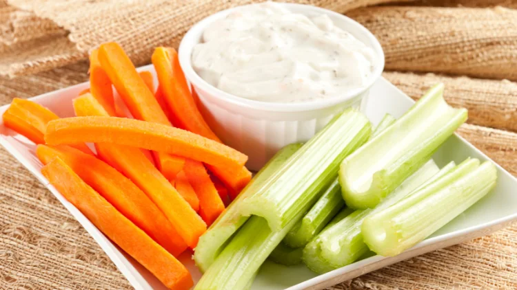 Slices of carrots and celery lay on a beige fabric alongside a small bowl of ranch dressing, all served on a white plate.