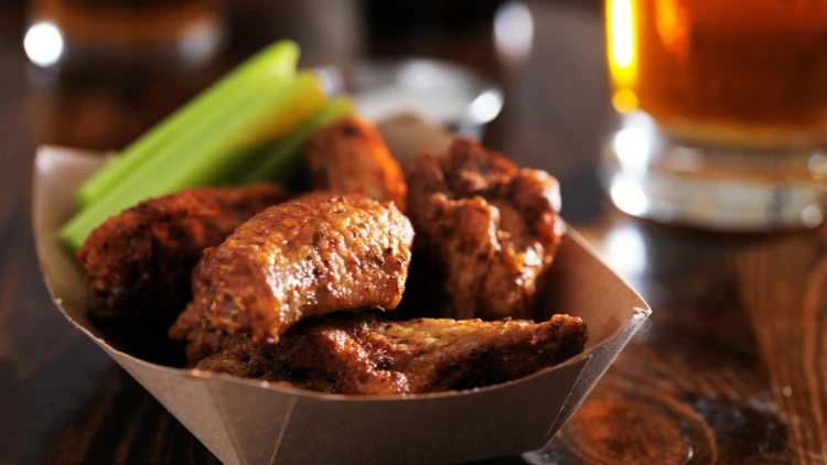 A to-go container with a few chicken wings with celery sticks displayed on top of a shiny wooden surface and a glass of beverage blurred in the background.