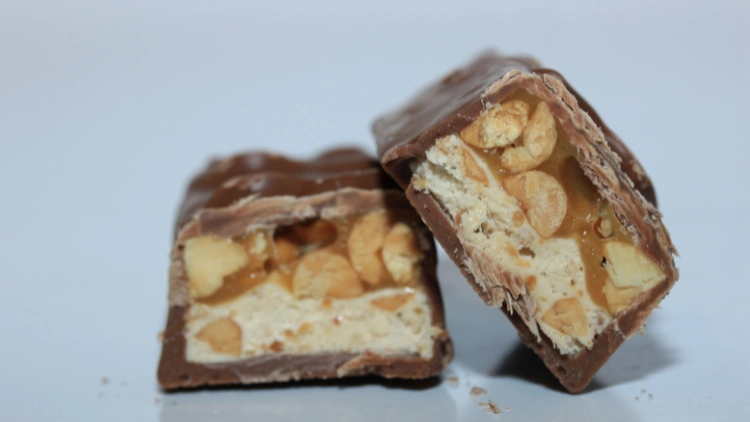 A bar of chocolate candy cut in half showing the inside filled with nuts and caramel, placed on top of a shiny white surface.