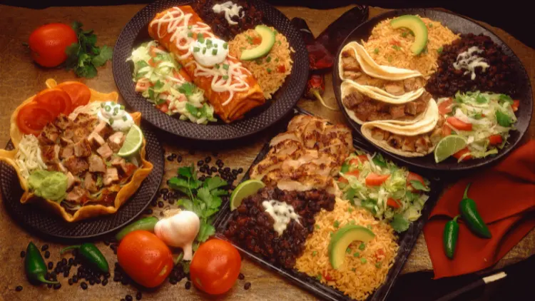 Different kinds of Mexican food that are made of ingredients such as chicken meat, sour cream, tomato, chili pepper, avocado, and other vegetables served on top of a wooden table.