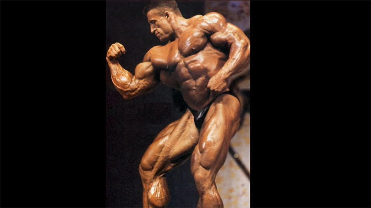 Dorian Yates, a professional bodybuilder is standing on stage flexing his bicep and pecs with striations seen in his entire body, although his upper chest is lagging. 