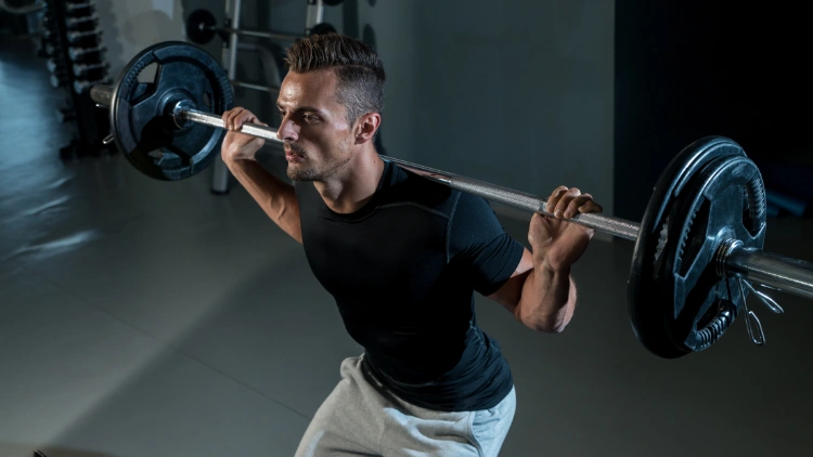 A fit man wearing a black activewear shirt, grey short is performing a barbell back squat using a barbell with two weighted plates in a dimly lit studio.
