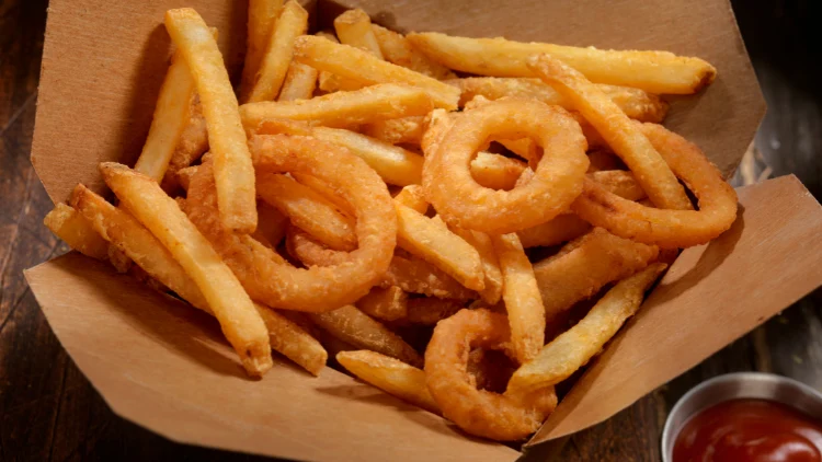 A to-go box filled with French fries and onion rings, and beside the box is a ketchup sauce, placed on top of a wooden surface.