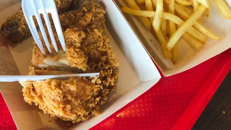 Two pieces of fried chicken, one is being sliced open and on the side are fries both in to-go boxes on a red tray.