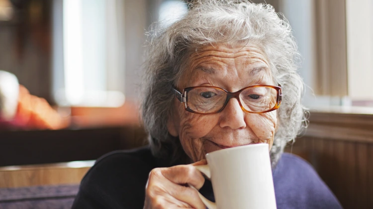 A Grandmother with eyeglasses, wearing a purple sweater, holding a white coffee cup full of black coffee.