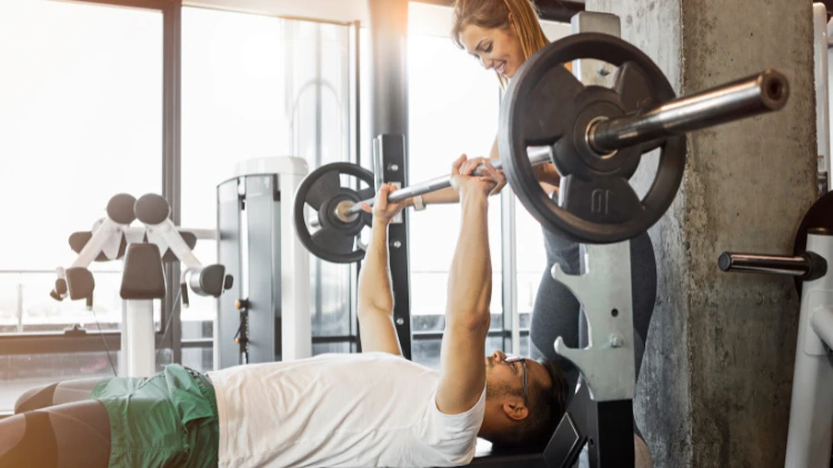 A man wearing a white t-shirt and green short is on a bench grabbing on to the barbell, setting up for a bench press exercise while a woman is assisting behind him.