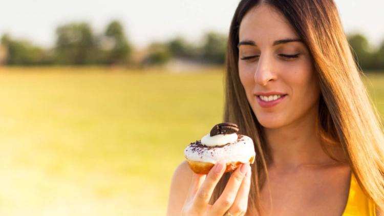 A woman wearing a yellow tank top, smiling while looking at her donut outdoors with a view of a field in the background.