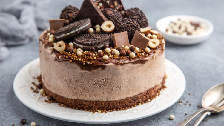 A chocolate ice cream cake topped with cookies and chocolate bits on top served on a white plate with spoon and fork on the side and displayed on top of a grey fabric.