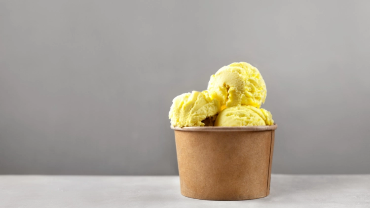 A yellow-colored ice cream scoops in a brown paper cup placed on top of a grey surface with grey wall in the background.