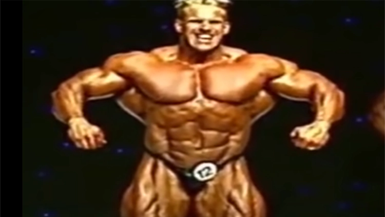 A blonde man is flexing his bicep and chest muscles which are toned and large as he has a large, smiling grunting face. 
