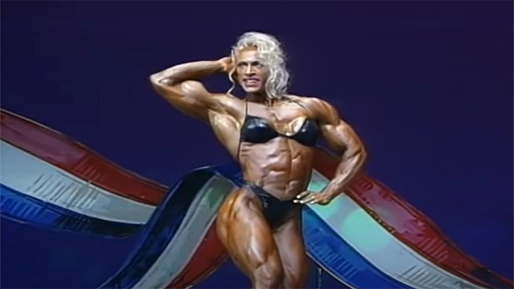 A blonde woman wearing a black bikini top and bottom is posing in front of a red white and blue striped background with one hand on her hip and the other behind her head as she smiles with a very muscular physique. 
