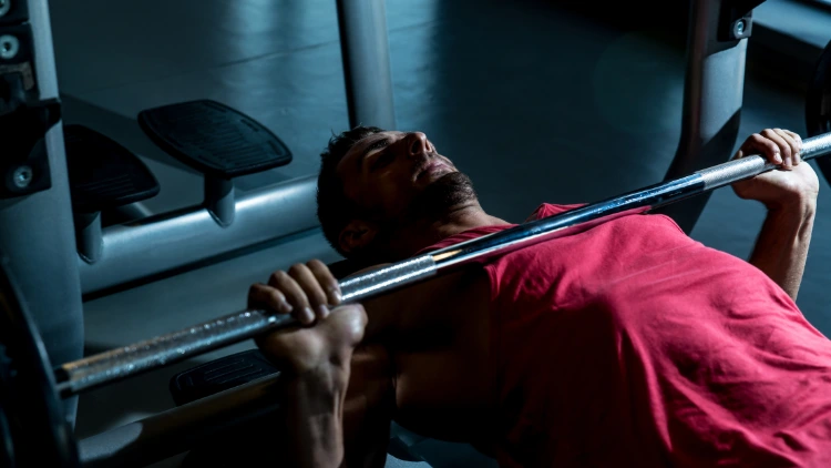 A man wearing a red tank top is in a dimly lit gym his laying down on a bench with his back arched and is in the starting position of a bench press. 