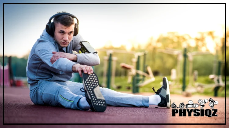 A man wearing large headphones, a phone arm band, grey pants and a grey hoodie is sitting on a track preparing for a morning run by bending over to touch his right shoe or toe to stretch his hamstring and there's playground equipment in the background. 