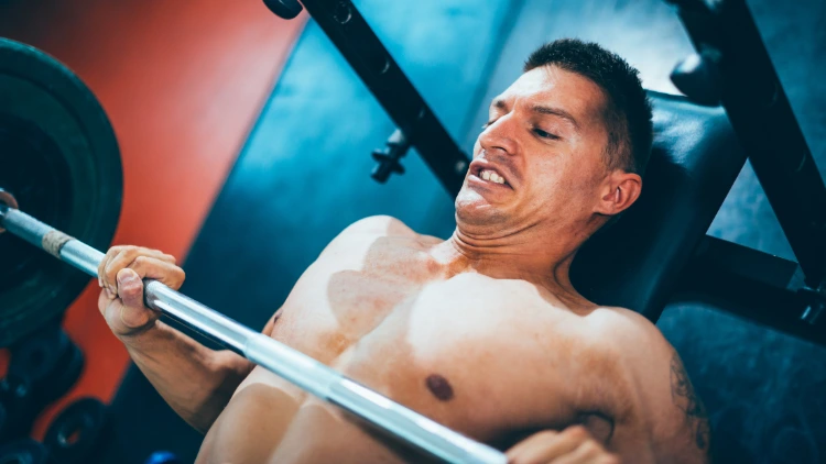A shirtless man with a struggling face is laying on a black colored bench in a gym holding onto a barbell too low almost touching his chest.