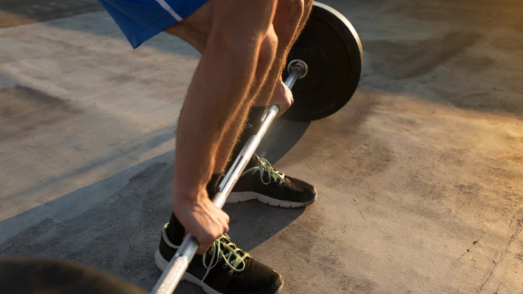 A man wearing a blue short, wearing black shoes is performing a light deadlift, using a barbell with only one weighted plate.