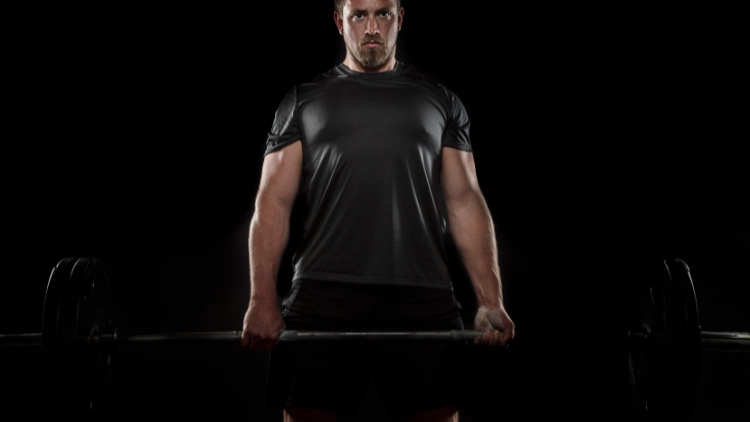 A man wearing a black activewear shirt and black shorts is holding onto a barbell with mixed-grip in a dimly lit studio.