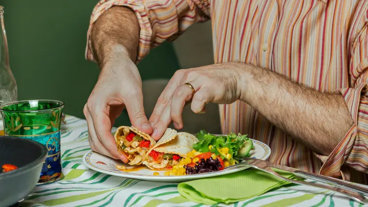 A man wearing a striped, orange long sleeves is sitting in a restaurant with green walls, as he checks inspects his plate with Mexican food such as tortillas.