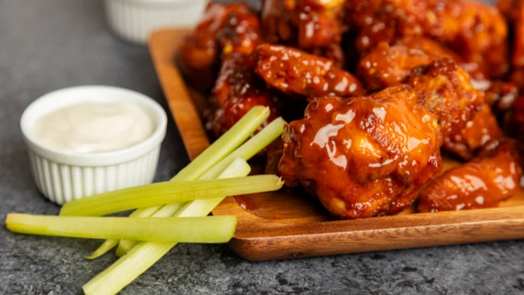 A few chicken wings in mango habanero variation served on a wooden plate with a cup of ranch dressing dip and five sticks of celery on the side.