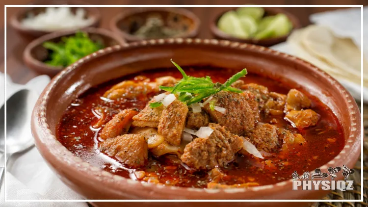 A bowl of menudo rojo with meat, green onions, white onions, tortillas and other garnishes on top with small bowls of the same garnishes in the background and blurred out. 