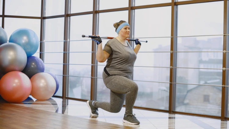 An overweight woman with blue headband and black gloves wearing a matching grey activewear shirt, pants and shoes doing a reverse lunge with a bar and exercise balls in red, yellow, blue and purple colors are visible in the background in a gym with large window with a view of foggy outdoors.