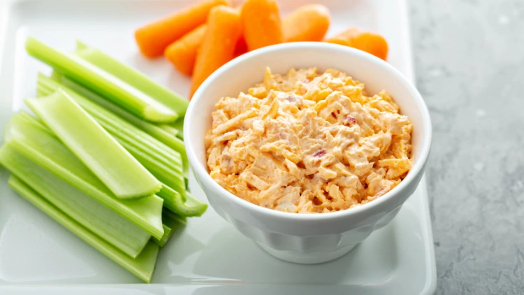 Pimento cheese on a small white bowl placed on a white square plate along with some celery and carrots.