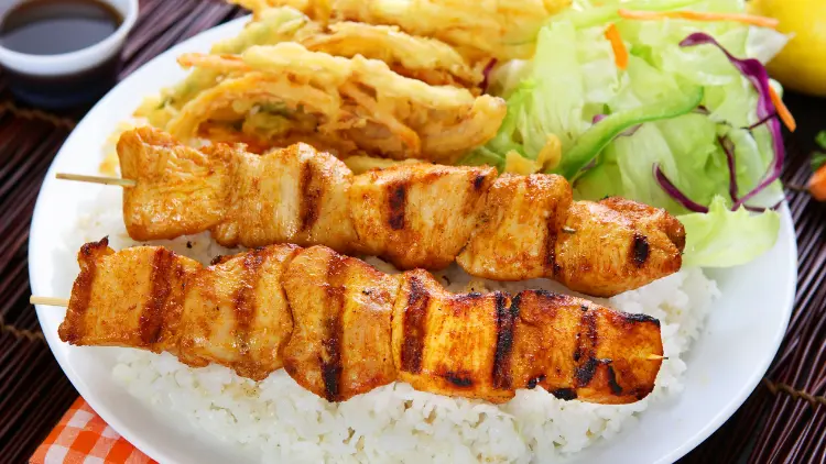 On a white dish, there's two sticks of chicken kabobs, rice, and cabbages, a small bowl of soy sauce can be seen in the background.