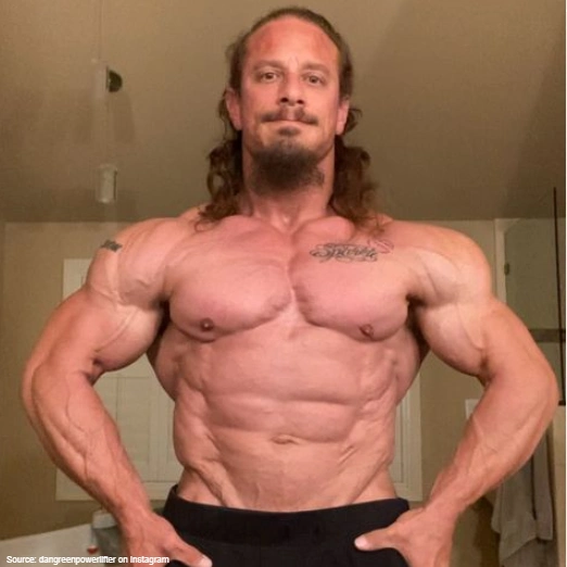 A powerlifter, Dan Green is posing in front of a mirror while lean, but his torso is blocky from focusing on compound movements and his V-taper isn't prominent. 