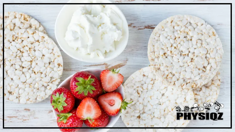 A white table has five plain rice cakes on top of that are also white in color and there's a bowl of cream cheese at the top and a cup of seven bright red strawberries at the bottom. 