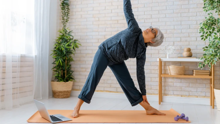A woman with short grey hair is standing in a yoga pose on an orange yoga mat with a laptop and a couple of purple dumbbells in the middle of a living room with a side table and a plant in the background.