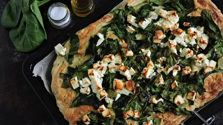 A rectangular-ish pizza topped with leafy vegetables such as spinach and placed on a black pan, beside it are bottles of seasonings and a leaf.