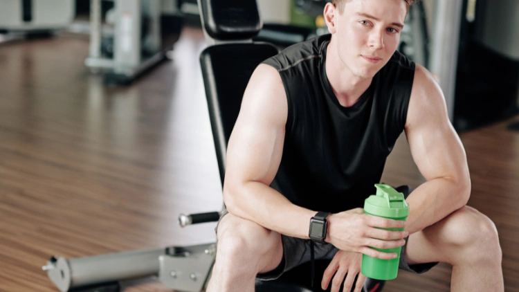 A man wearing a black tank top and shorts is seating on an inclined bench while holding a green water bottle filled with vitamin water, in a gym with wooden floor.