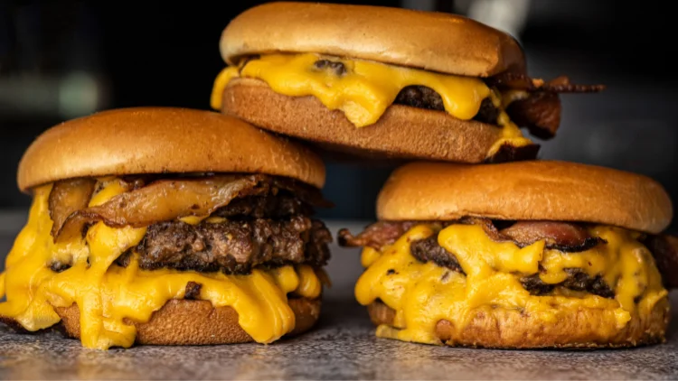 On top of a marble surface is a stack of three burgers with bacon and cheese dripping off the side.