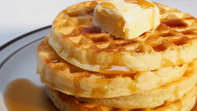 On a white dish is a stack of four pieces of waffles with butter on top and drizzled with syrup.