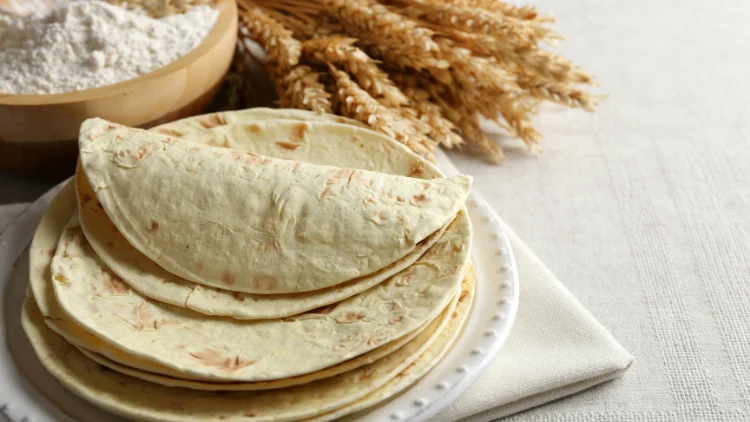 A pile of tortillas with one folded tortilla at the very top, placed on a white plate and displayed on top of a white fabric on a table with whole wheat and flour in the background.