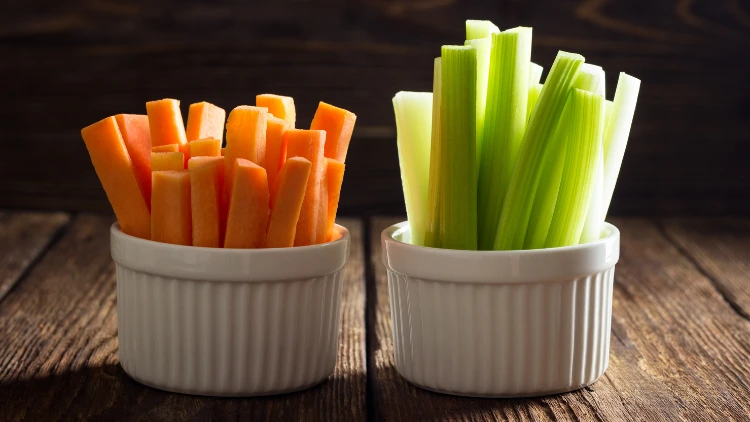 A few slices of carrots and celery served in separate white cups and displayed on top of a wooden surface.