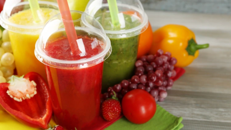Three clear plastic cups of vegetarian smoothie, one is in red-colored smoothie which can be a tomato, pepper or strawberry flavor, another cup has green-colored smoothie, and the other one has yellow-colored smoothie, displayed on top of a wooden surface.