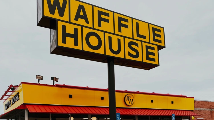A black and yellow post signage that says Waffle House and behind it is the Waffle House store with the logo WH.