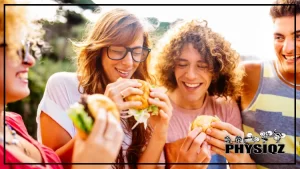 There's four people outdoors where two of them are in pink shirts and glasses, but the other two are in white and grey shirts and three of them are eating burgers but the man at the far right is just watches them eat while wondering if there's Whataburger gluten free options to be safe.