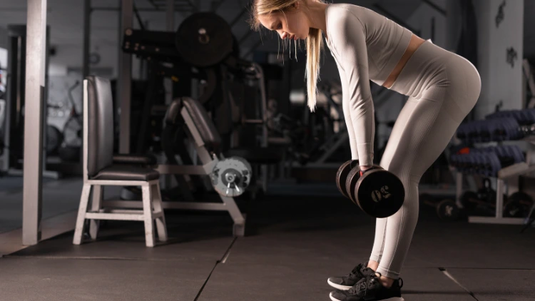 A blonde woman wearing a matching grey sweatshirt and leggings, and black shoes is in a gym holding two dumbbells as she does a stiff legged dead lift by hinging at the hips with her back straight and legs extended with a slight bend at the knees while her arms are hanging in front of her.