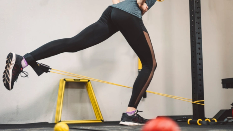 A woman wearing black leggings is standing on her right leg while her left leg is extended behind her in a glute kick back, her hips are at a slight hinge, and has her ankle attached to a cable.