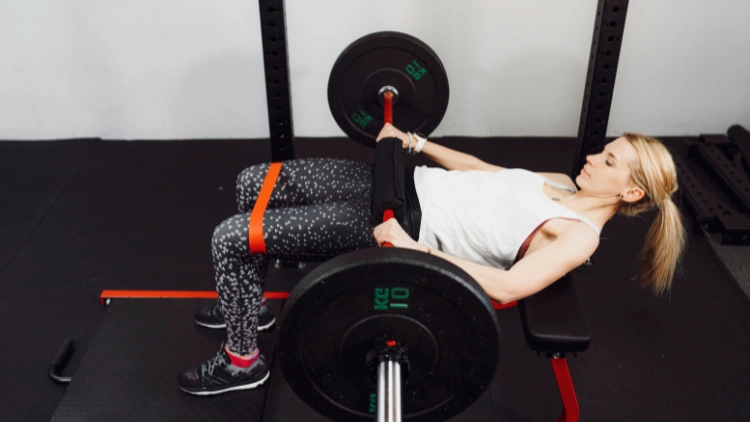 A blonde woman wearing a white tank top and a dotted-pattern black leggings, and black shoes is in a gym laying with her upper back on a bench with her chest and back extended over the end, knees bent at a 90 degree angle while holding an empty barbell at her waist as she does a hip thrust. 