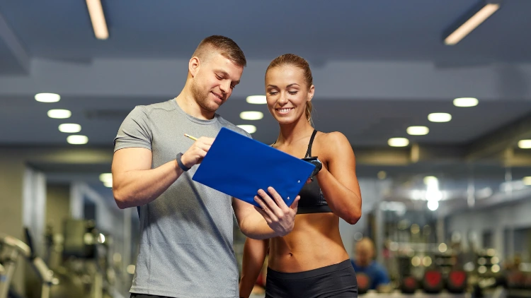A woman wearing a black sports bra and shorts is looking at the workout plan that her personal trainer made who is beside her wearing a gray activewear shirt and black short in a gym.