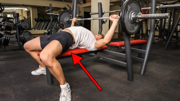 A guy in a white tank top is holding a barbell at the top of a bench press with his lower back overly arched which makes his glutes rise off the bench. 