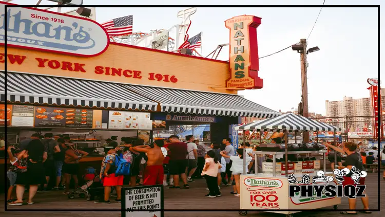 Several people are lining up at a hotdog place that has store signage that says, "Nathan's famous frankfurters" with the lettering-colored yellow, red and green, some of the customers are wondering, "are Nathan's hot dogs keto friendly", as they order their food from the store and the food stall nearby.