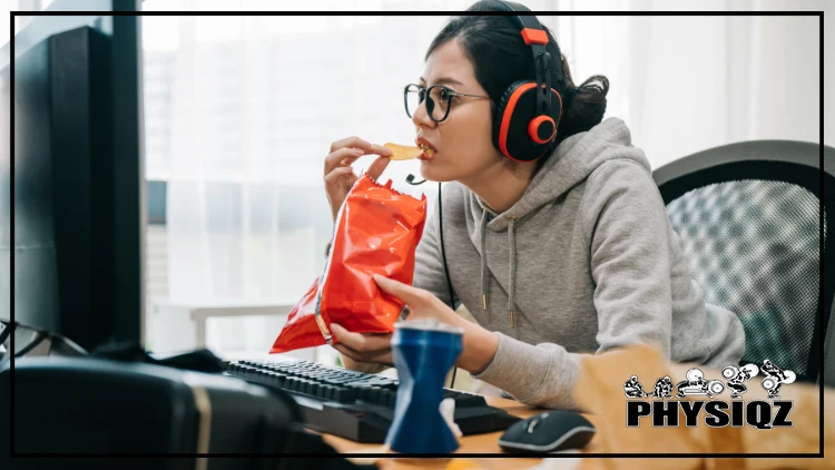 A woman with glasses and headphones on, wearing a grey hoodie, at home sitting in front of her computer browsing for an answer to her question "are Quest chips keto" while eating snack from a red bag of chips.