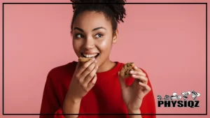 A young woman wearing a red sweater gives a curious face wondering "are Quest cookies keto", while taking a bite from a piece of cookie and holding another piece of cookie on the other hand.