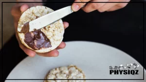 A person wearing black clothes is wondering "Are rice cakes keto" as he spreads chocolate filling on a rice cake with both hands while sitting in his room with another piece of rice cake in front of him on a white plate.
