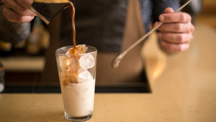A barista is shown pouring espresso over a glass cup filled with ice and milk, while holding a long spoon in his other hand for stirring, the espresso is dark and rich, and is being poured from a small glass pitcher with a spout, the glass cup is partially filled with ice and milk.
