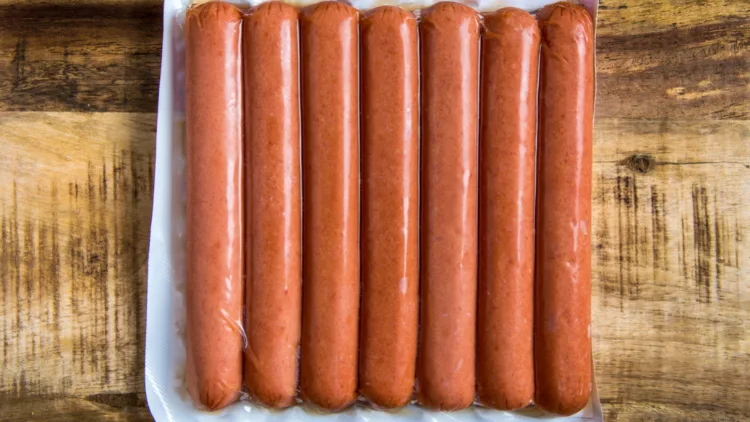 Seven pieces of raw beef frank hotdogs sealed tightly in a plastic bag and placed on top of a wooden board.