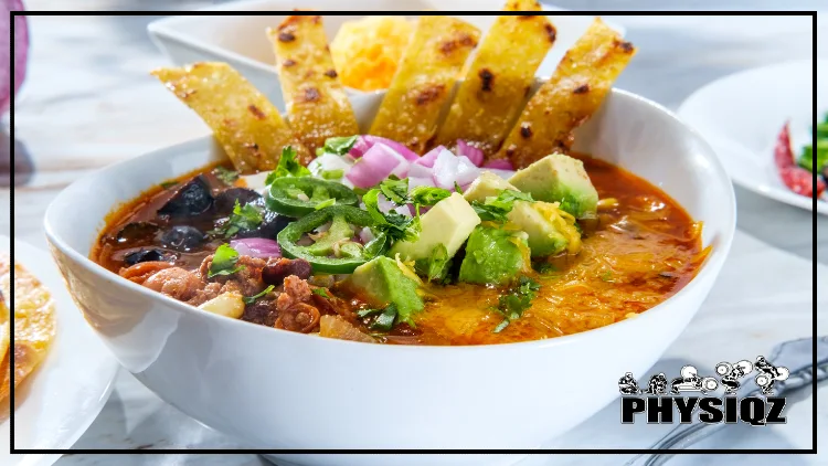 A white bowl is filled with soup including the ingredients broth, avocado, black beans, jalapeno, red onion and tortilla strips. 